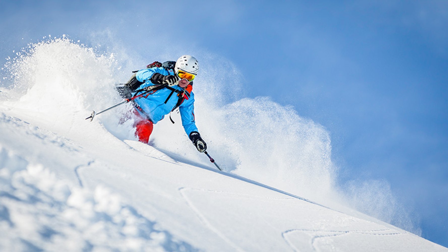 Mann på ski kjører ned en bratt bakke i puddersnø, med snøsprut rundt seg under klar blå himmel.