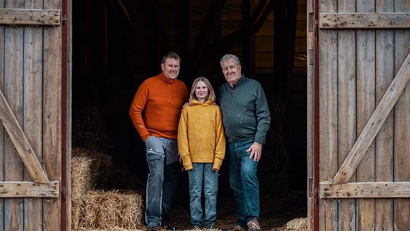 Tre generasjoner menn står i døråpningen til en låve.