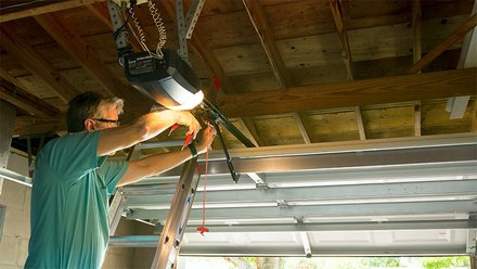 Person står på stige og utfører arbeid på en garasjeportåpner montert i taket.