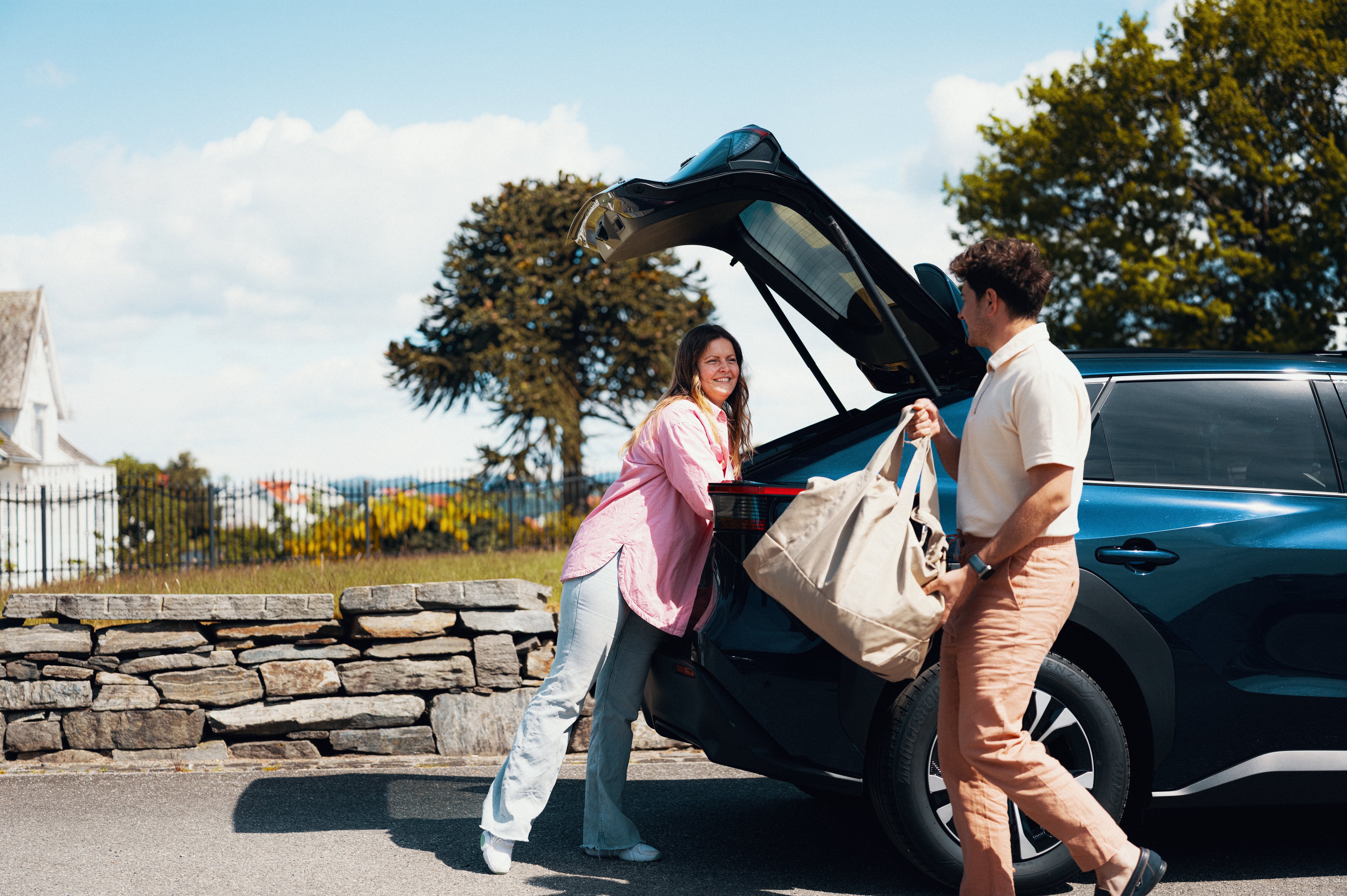 En smilende dame og en mann legger baggasje i baggasjerommet på bilen.