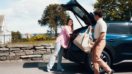 En smilende dame og en mann legger baggasje i baggasjerommet på bilen.