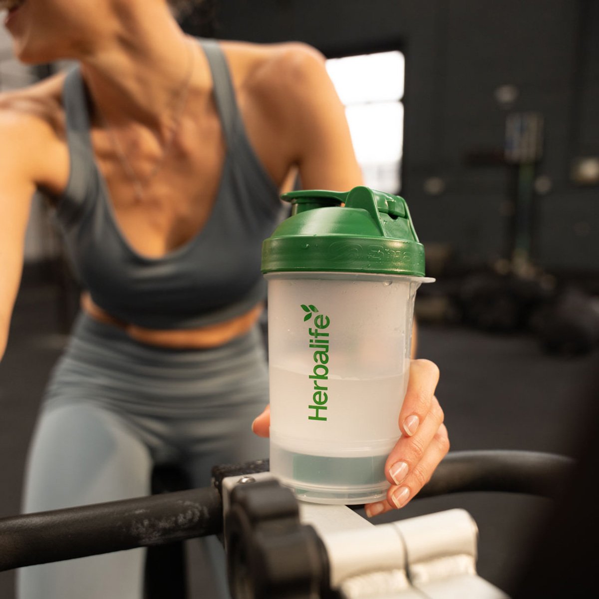 Donna in palestra tiene uno shaker con il logo Herbalife vicino a un attrezzo