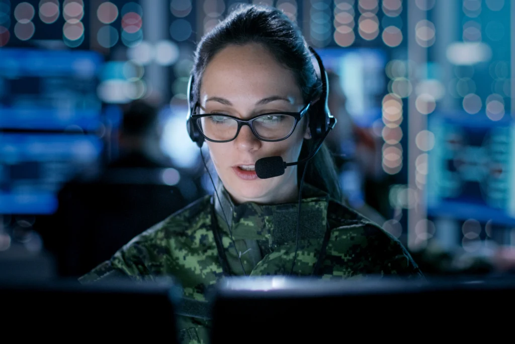 Person wearing a headset and glasses working at computer monitors in a control room, with blurred server racks and network equipment in the background. Person wearing a headset and glasses working at computer monitors in a control room, with blurred server racks and network equipment in the background.