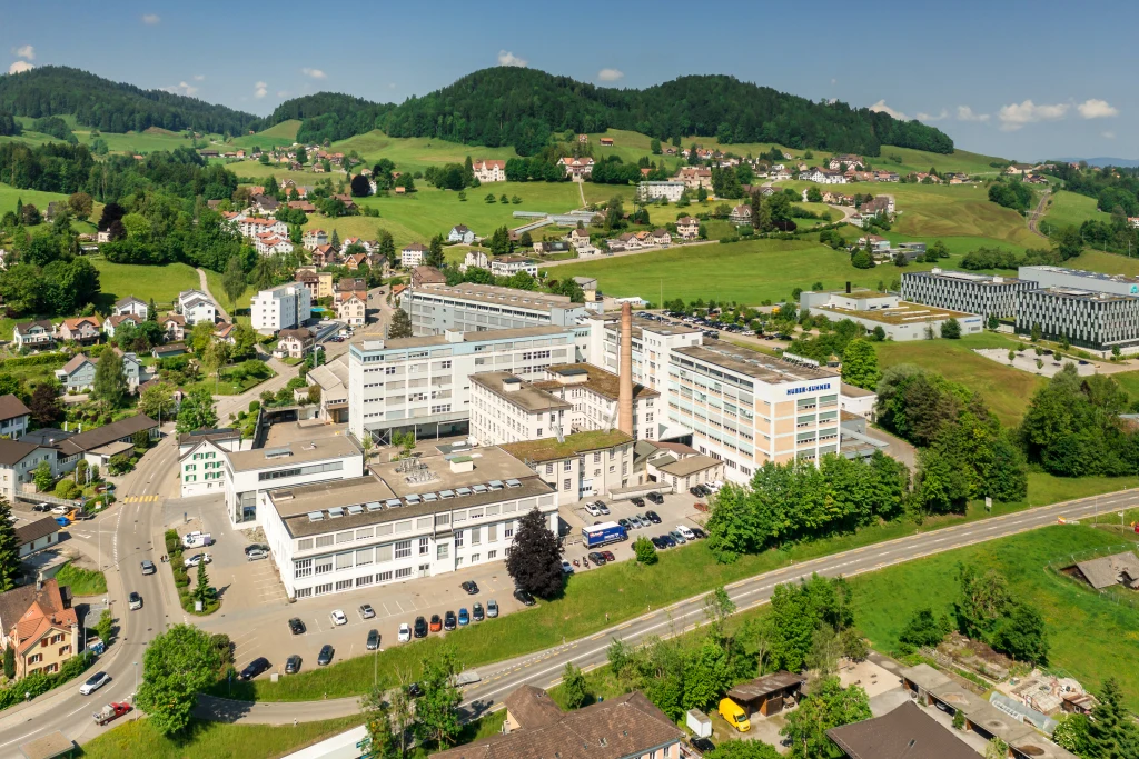 Aerial view of HUBER+SUHNER’s Herisau site in Switzerland, surrounded by green hills, farmland, residential buildings and roads.