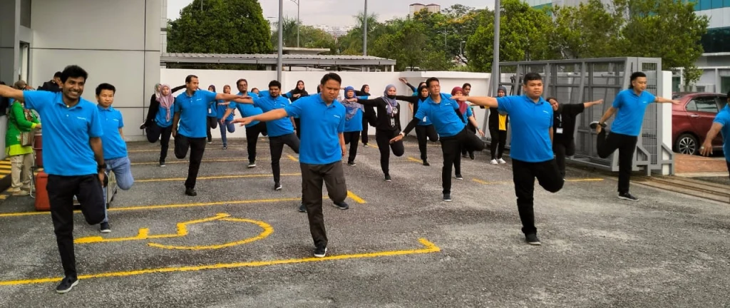 Körperliche Aktivitäten werden gefördert – auch während der Arbeit: SenamRobiks ist eine Aerobic-ähnliche Sportart, die unsere Mitarbeitenden seit zehn Jahren praktizieren Mitarbeitende machen Dehnübungen auf dem Parkplatz des Büros in Malaysia.