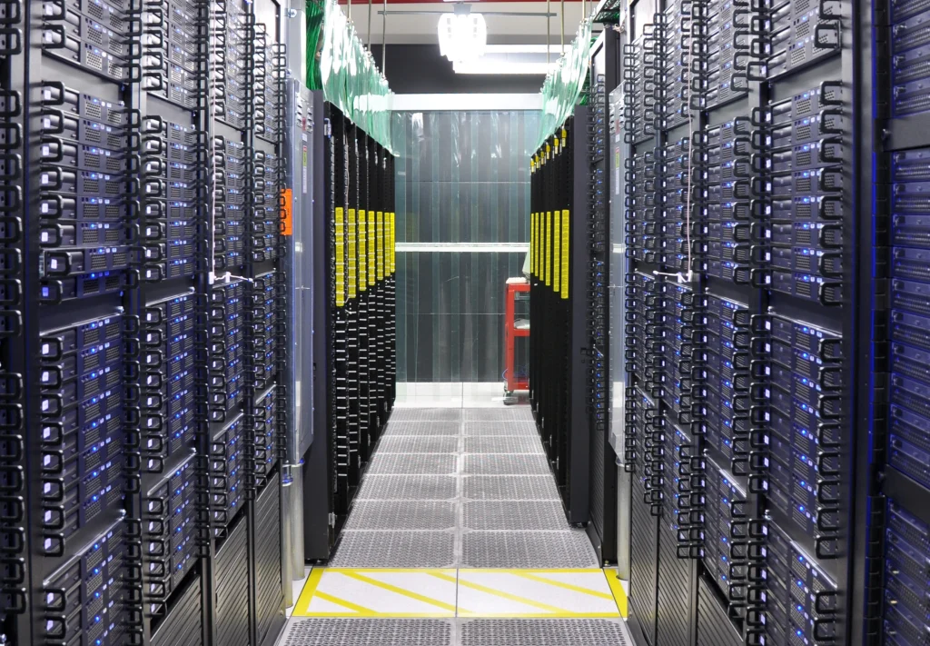 Data center aisle lined with server racks on both sides, blue status lights glowing, under bright overhead lighting in a secure facility.