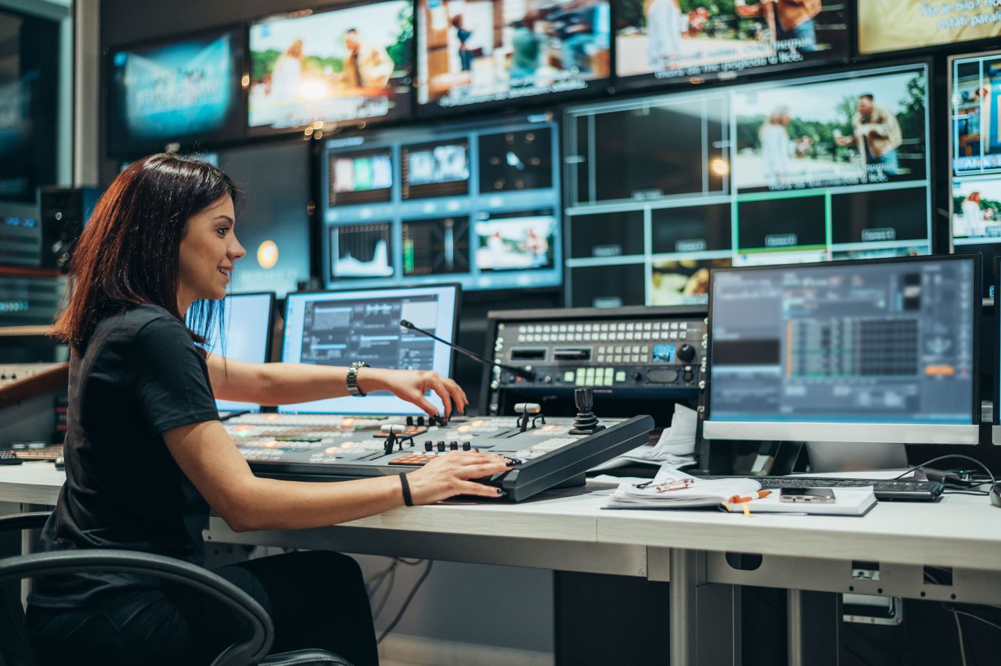 A lady operating a video broadcast control panel with multiple screens displaying live footage in a media production studio.