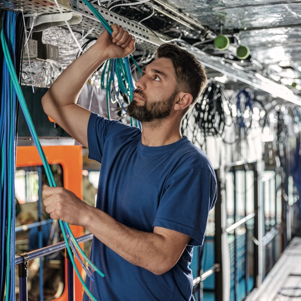 Ein Techniker in einem blauen Hemd verlegt und verbindet Kabel im Inneren eines Fahrzeugs, umgeben von Leitungen, Kabeltrassen und elektrischer Infrastruktur.