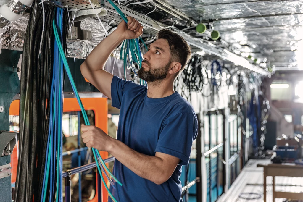 Techniker in blauem Hemd verlegt und verbindet Kabel im Fahrzeuginneren, umgeben von Leitungen, Kabeltrassen und elektrischer Infrastruktur.