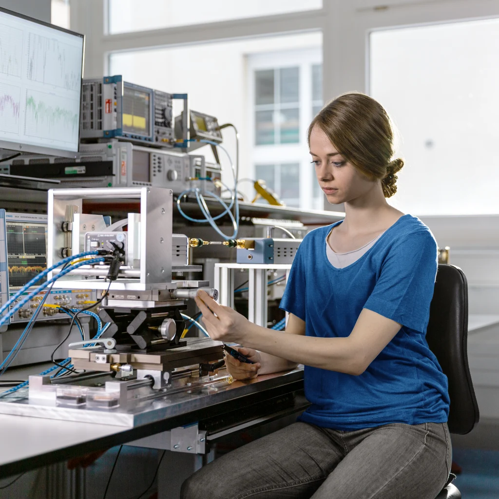 Eine junge Frau arbeitet in einem modernen Elektroniklabor an einem komplexen Testaufbau, umgeben von Messbildschirmen, Präzisionsgeräten und Kabelverbindungen.