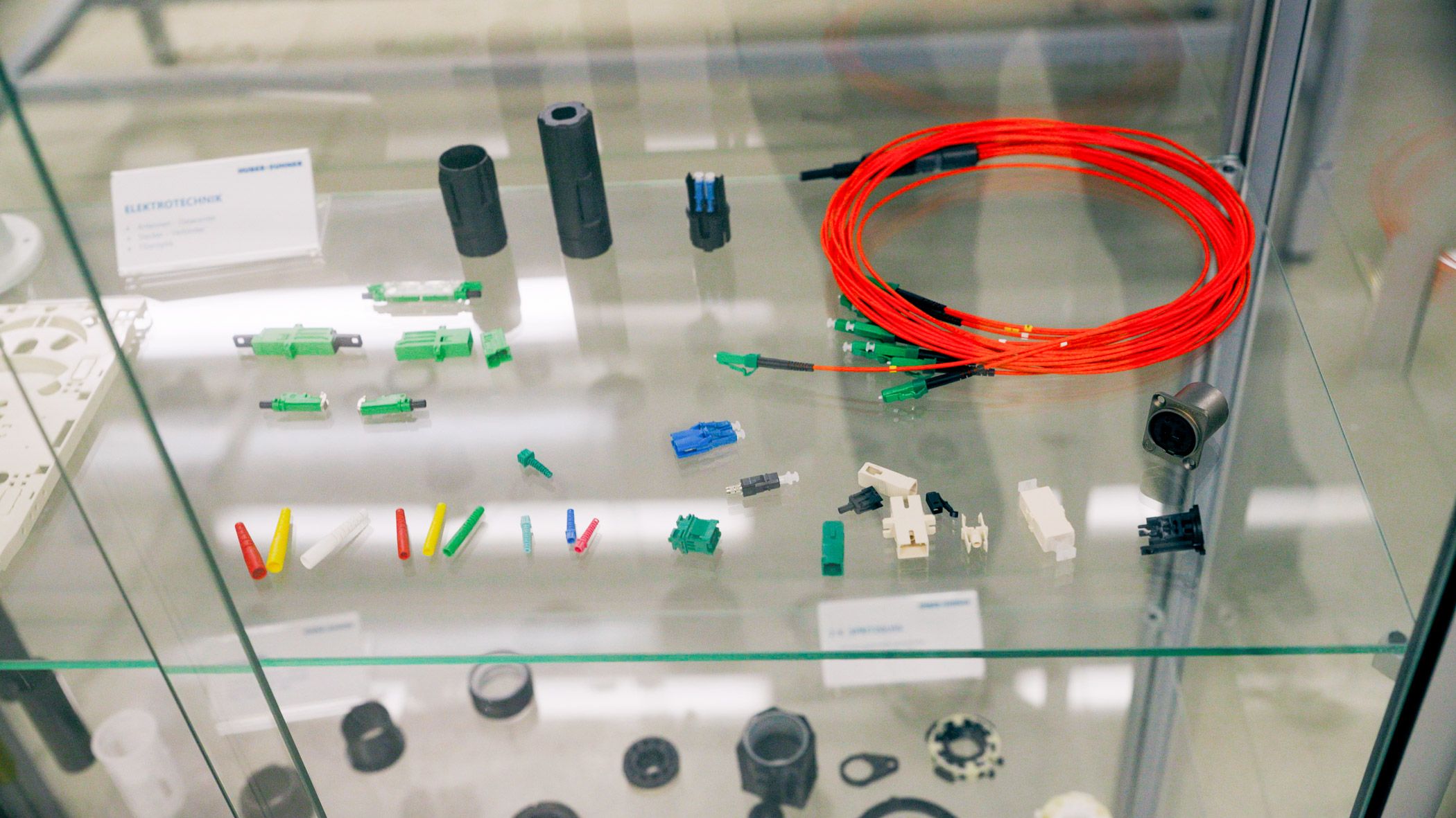 Various fiber optic connectors, adapters, and an orange fiber optic cable assembly displayed in a glass cabinet.