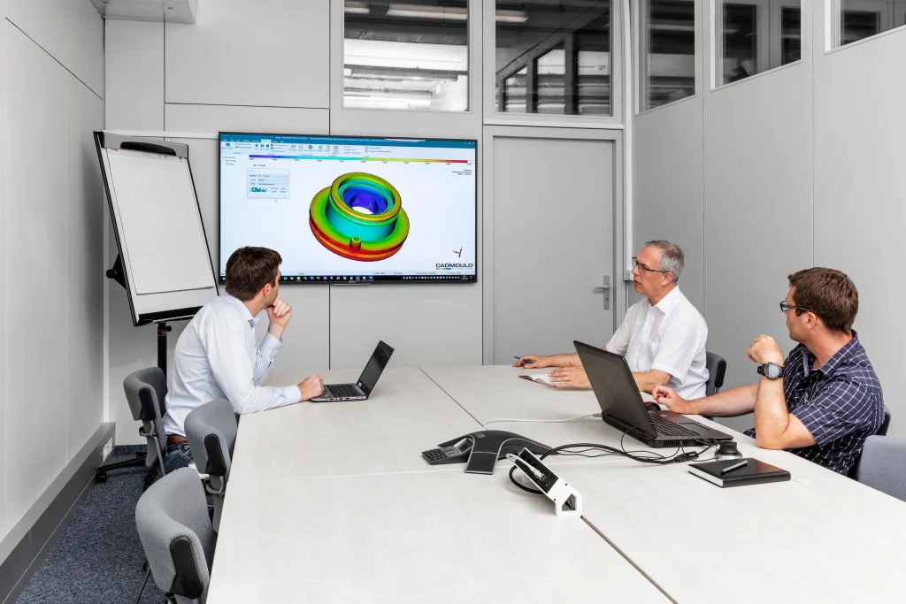 Conference room with three people using laptops while reviewing a colorful 3D CAD model displayed on a large wall-mounted monitor.