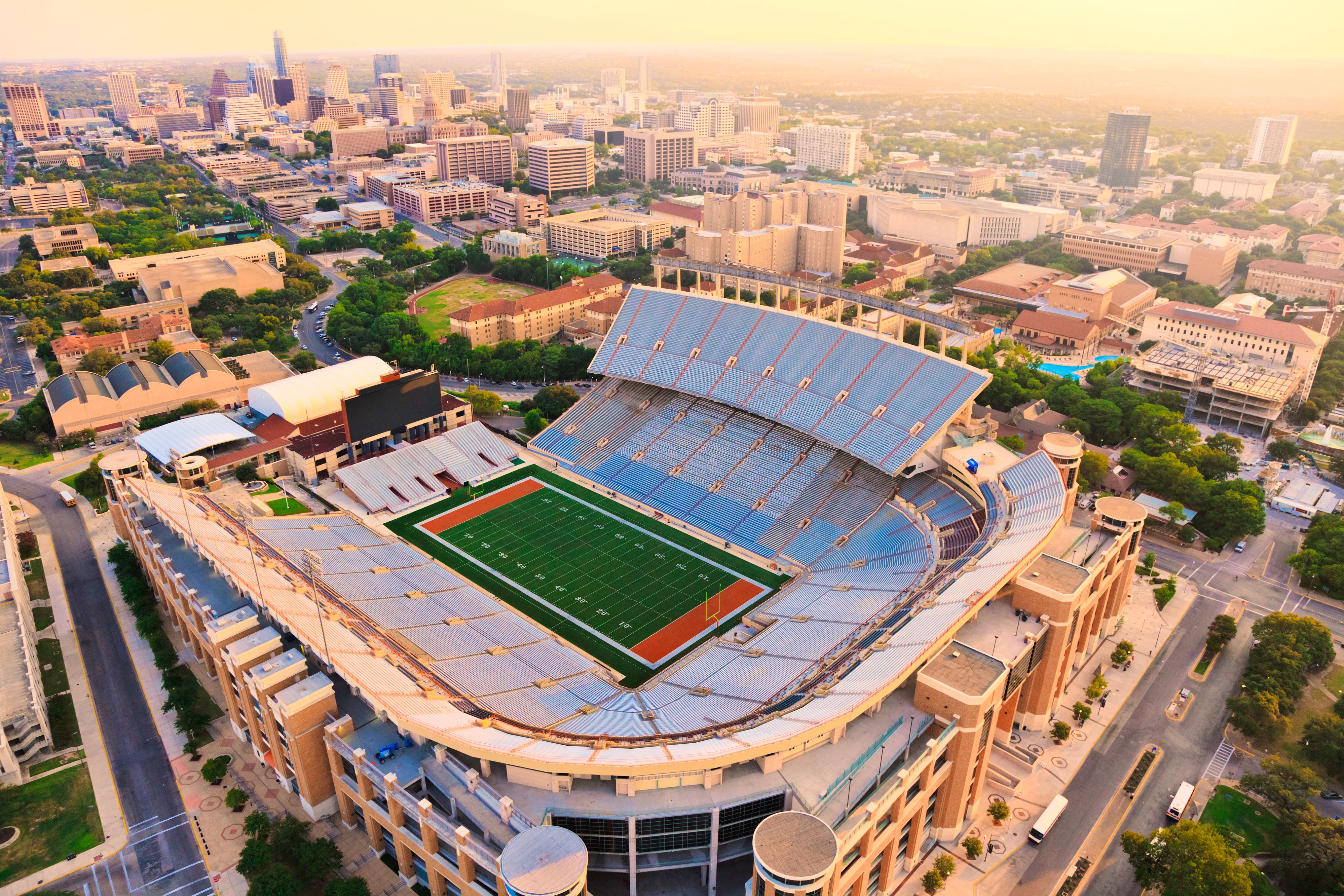 Aerial view of a large football stadium surrounded by a cityscape, with empty stands and a green field at the center.