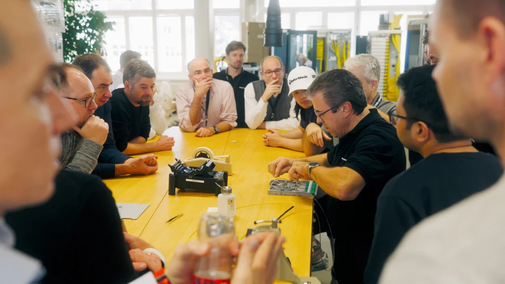 Mehrere Männer stehen um einen Tisch herum und beobachten einen Mann bei der Arbeit an einer Kassette.