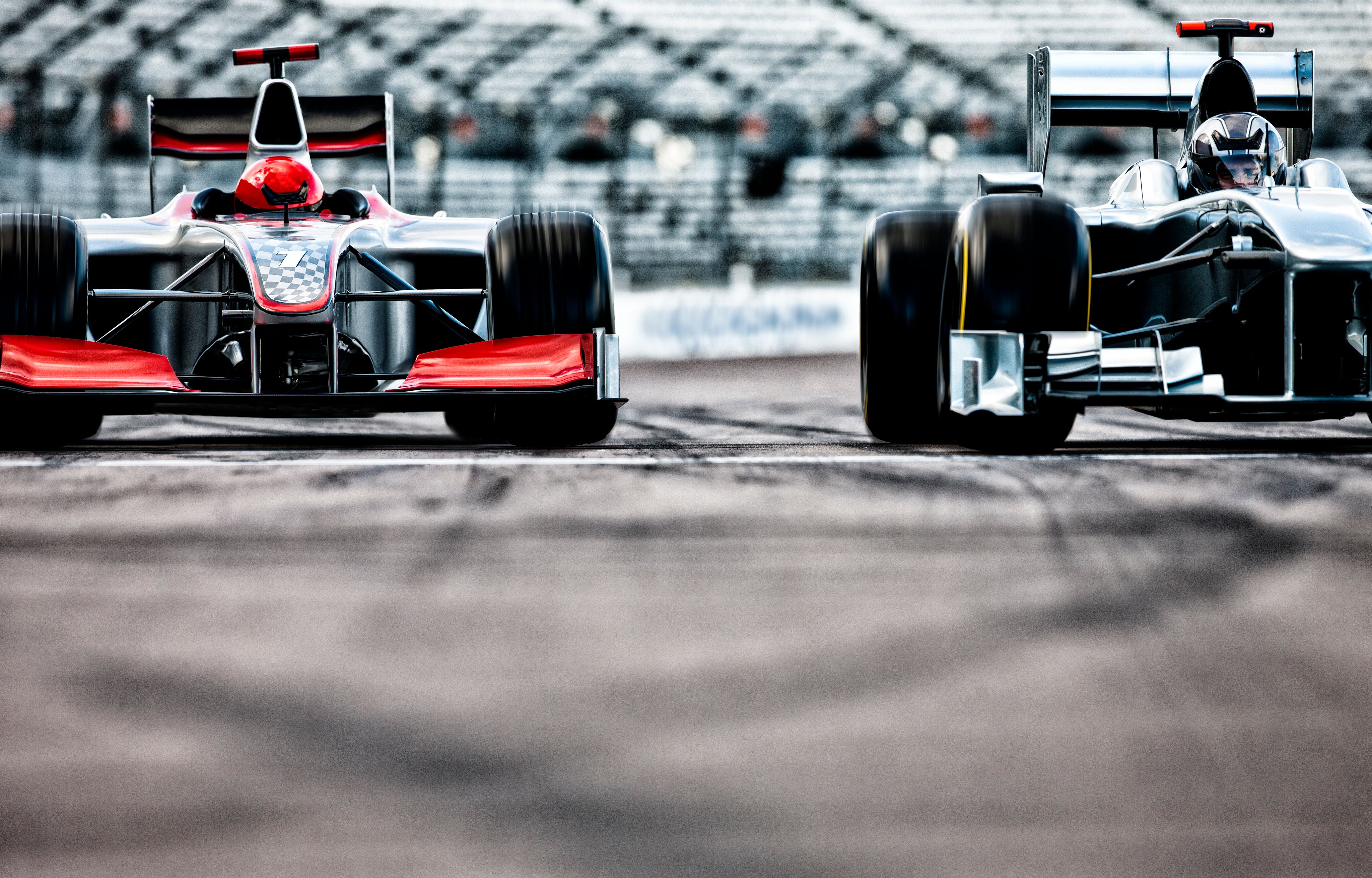 Two Formula-style race cars positioned side by side on a track, viewed from the front with blurred grandstand in the background.