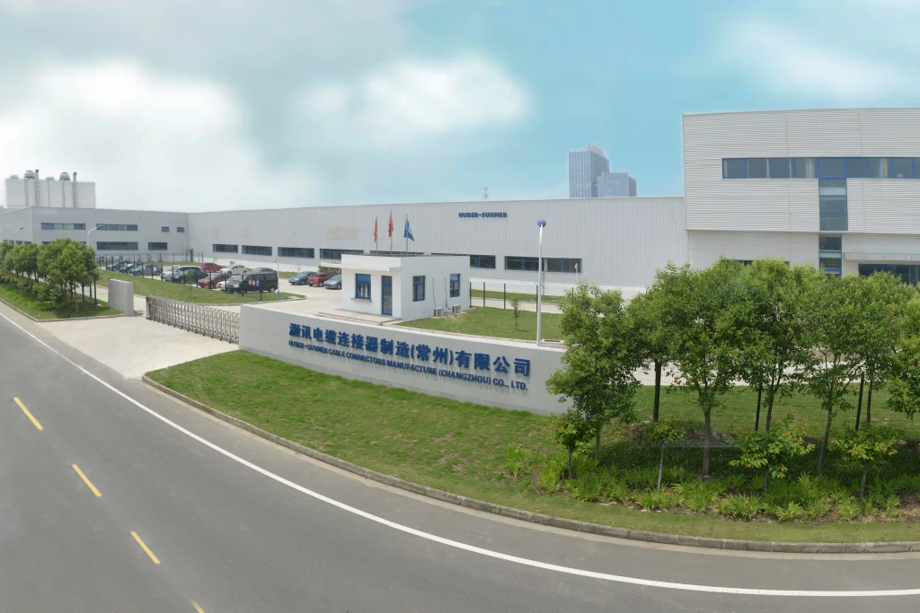 Modern industrial facility with white buildings, gated entrance, guardhouse, parked cars, and sign for HUBER+SUHNER Changzhou under a blue sky.