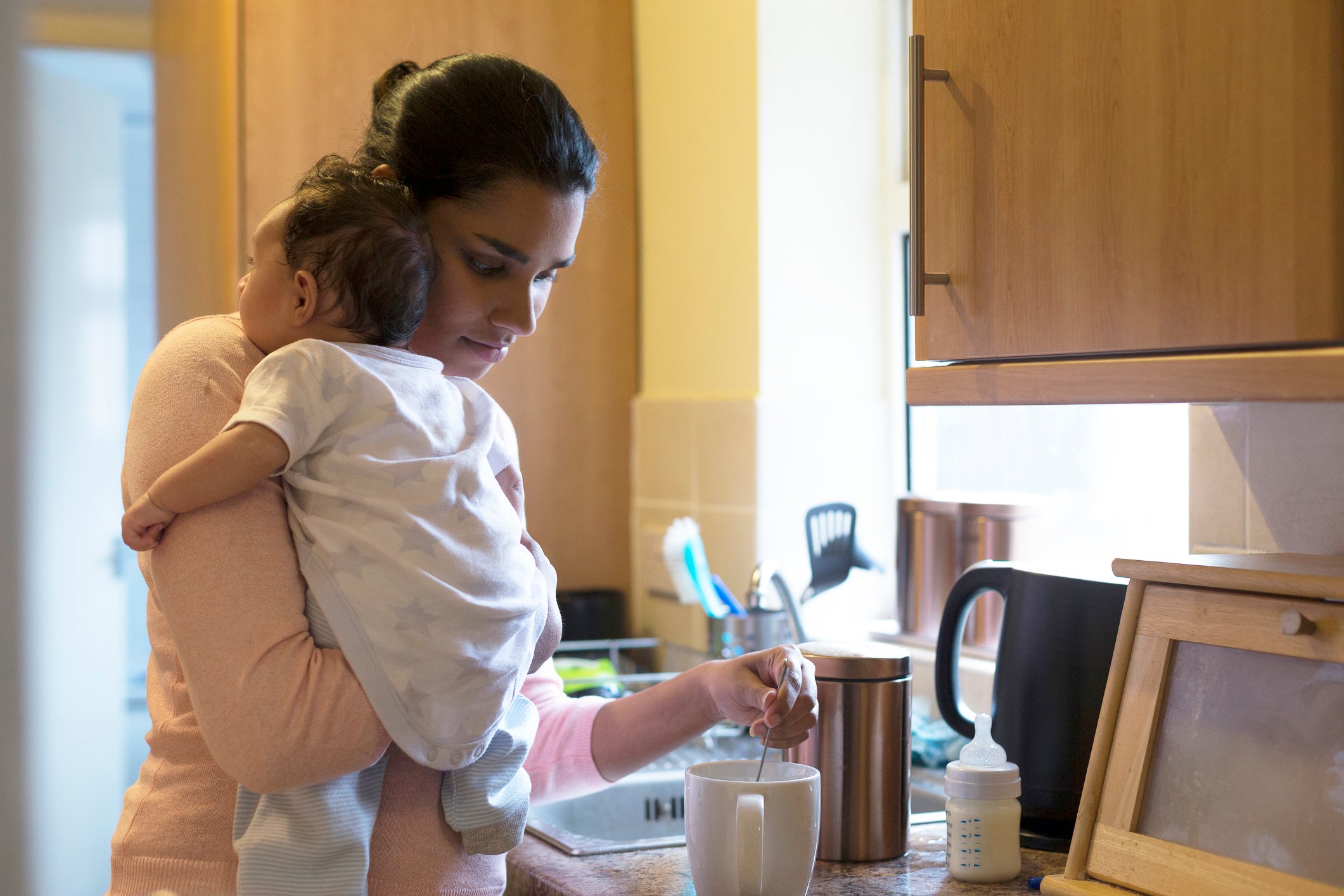 A mother holding a baby and making a cup of tea in a kitchen.