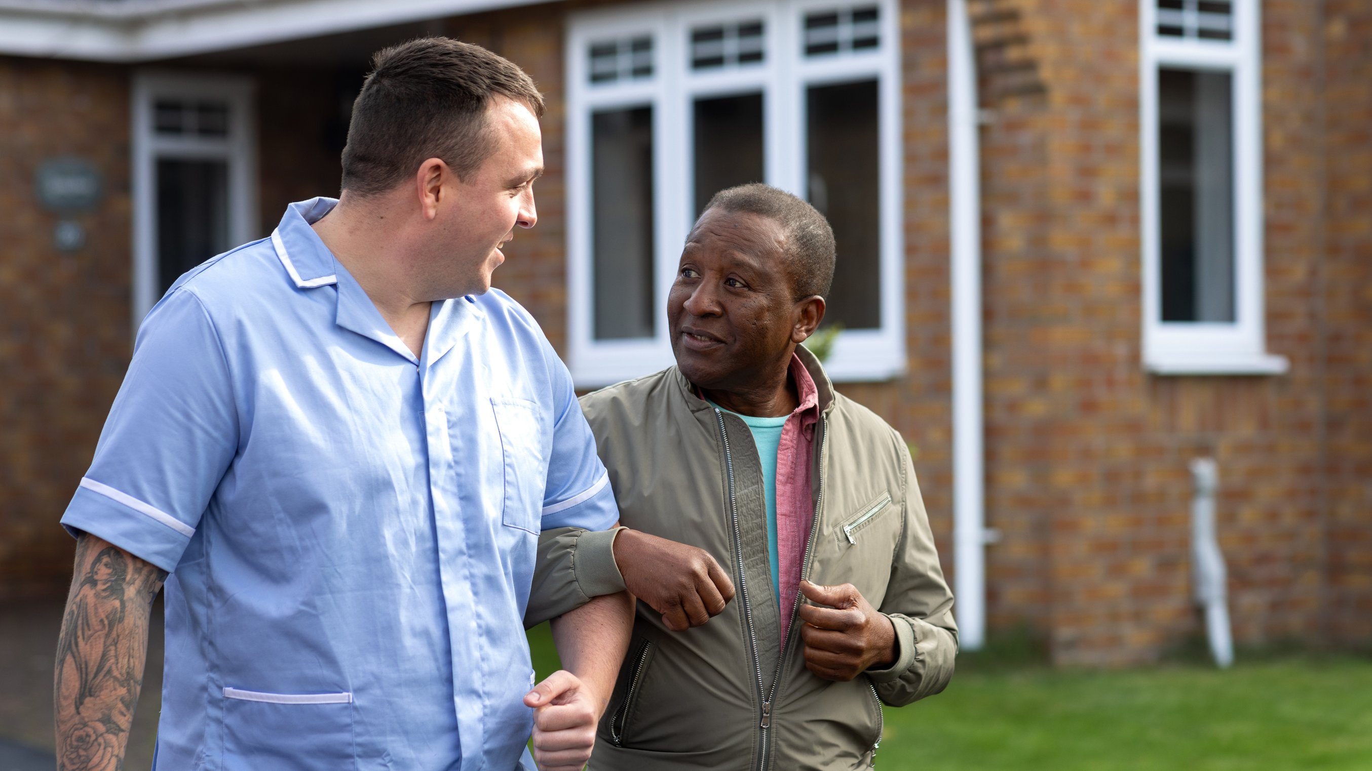 A care worker walking arm and arm with a mid life man who is being cared for outside a home.