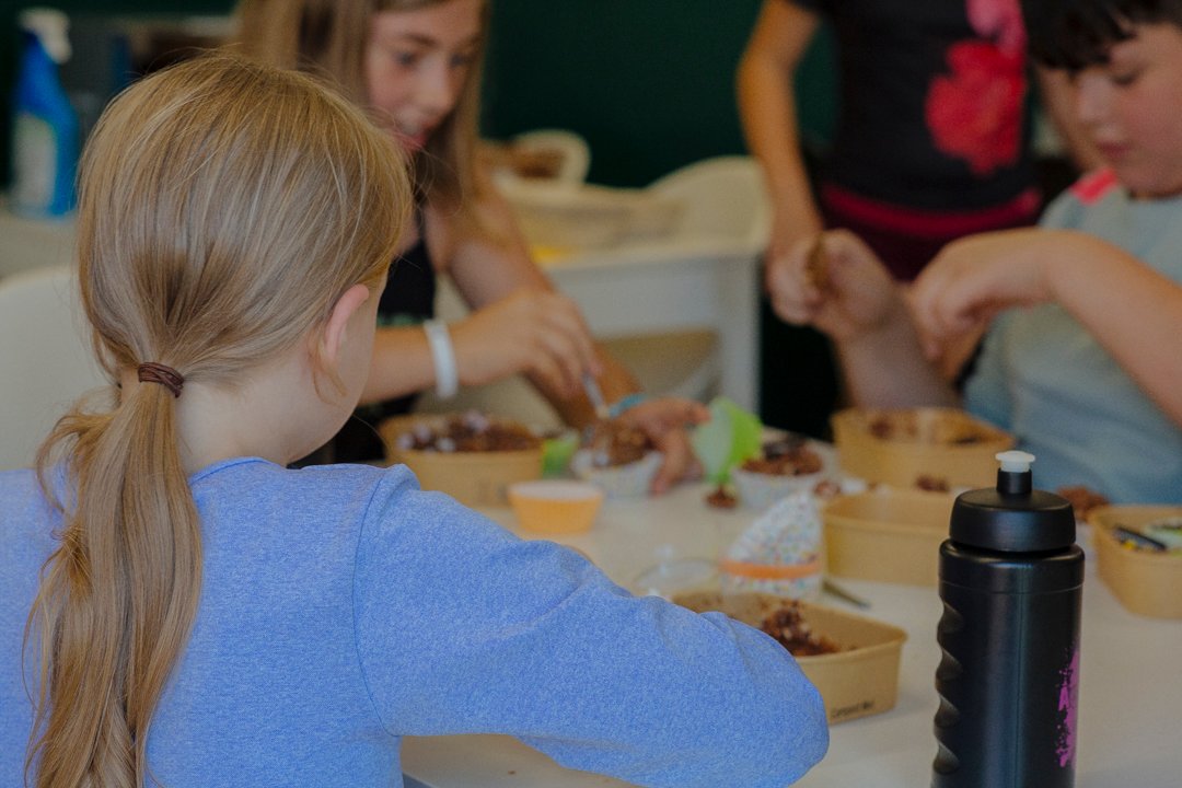 Three kids sat at a table making chocolate cornflake cakes at a community centre.