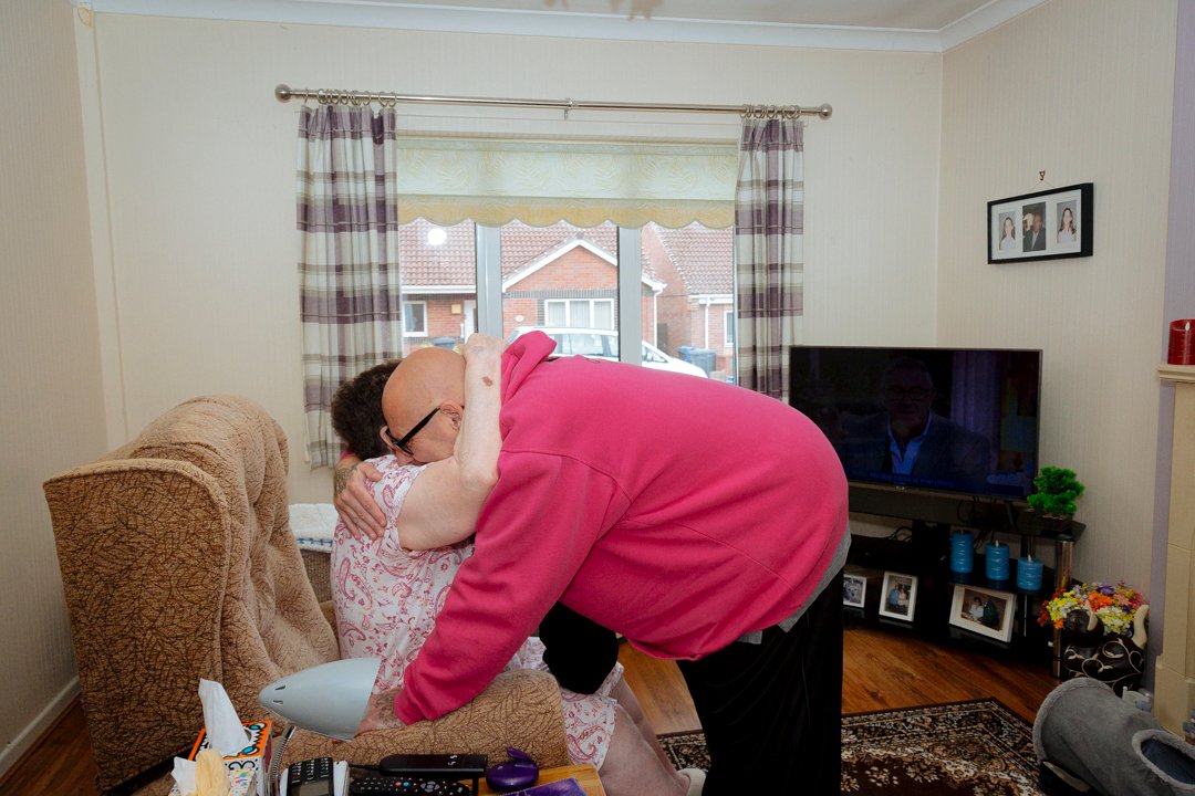 Elder person sat giving hug to care worker who is leaning down in their home.