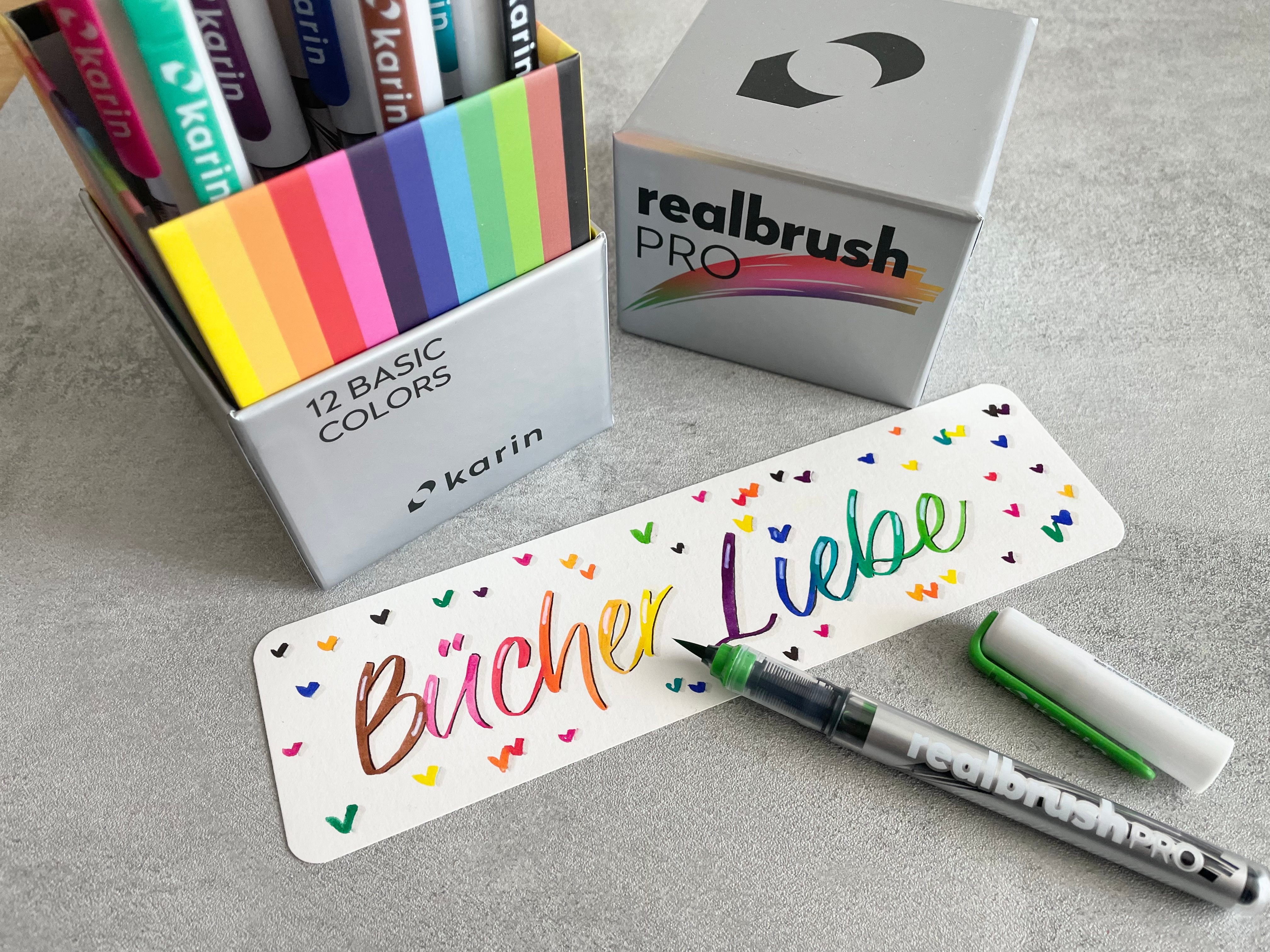 Fünf verschiedene Brush Pens auf weissem Schreibtisch mit Handlettering-Schriftzug, arrangiert neben einem offenen Notizbuch