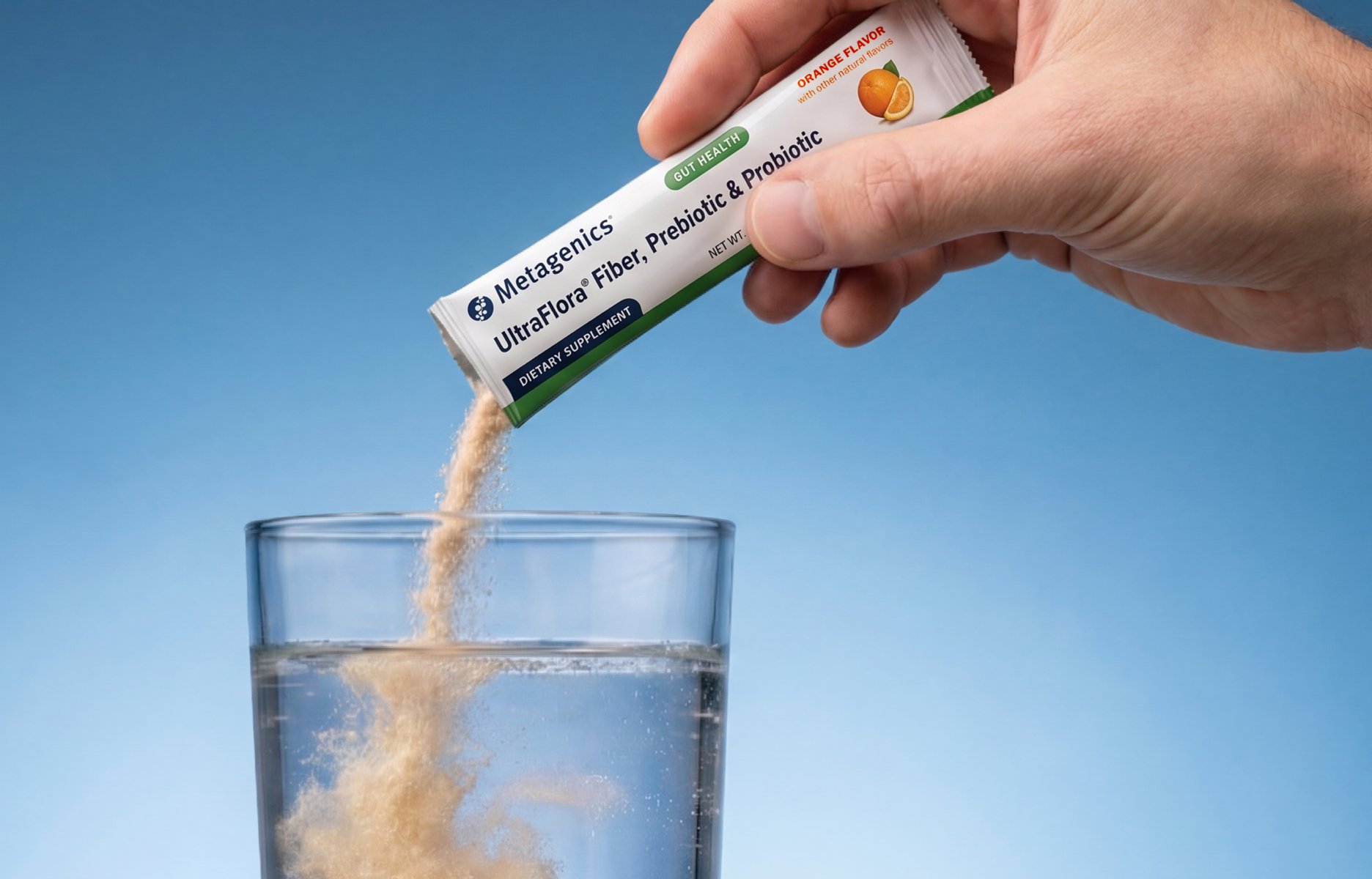 Hand pouring Metagenics' UltraFlora Fiber + prebiotics and probiotic, and fiber supplement powder into a glass of water against a light blue gradient background.