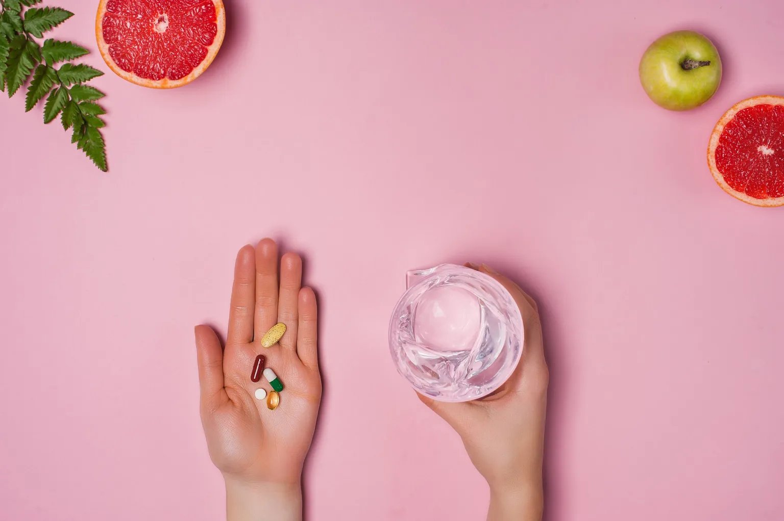 Hand holding several supplements beside a glass of water on a pink background with fresh fruit