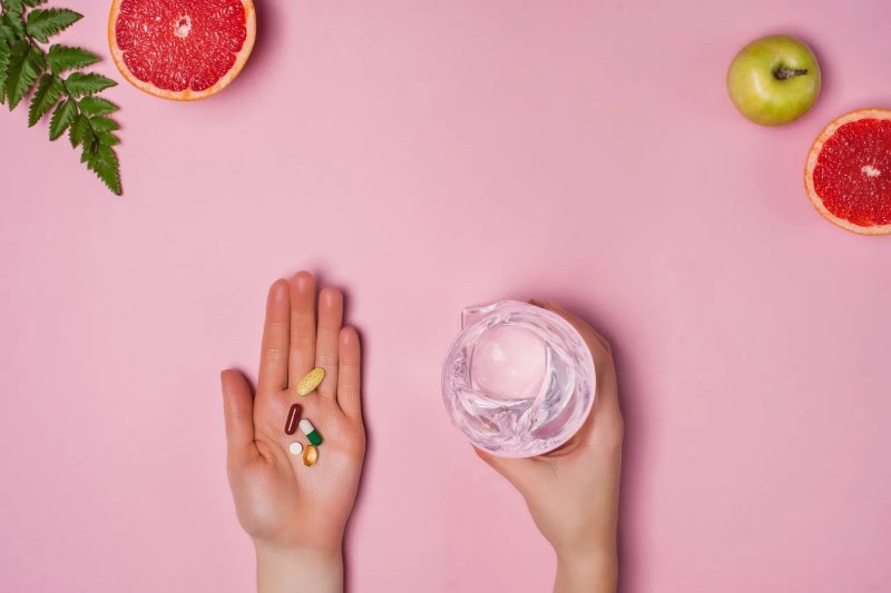 Hand holding several supplements beside a glass of water on a pink background with fresh fruit