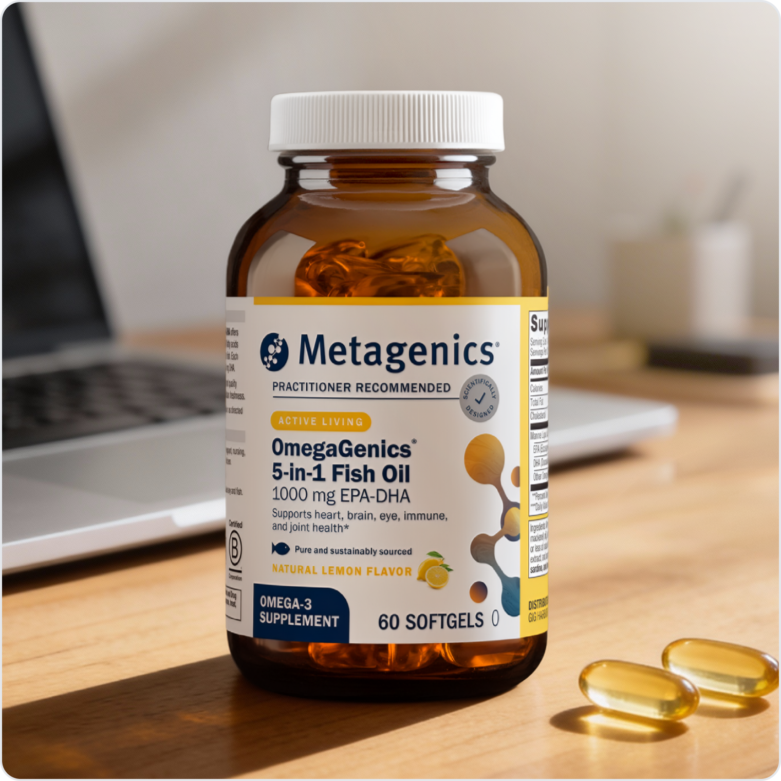 A bottle of Metagenics OmegaGenics® 5‑in‑1 Fish Oil 1000 mg EPA‑DHA on a wooden surface beside a laptop and several yellow softgel capsules.