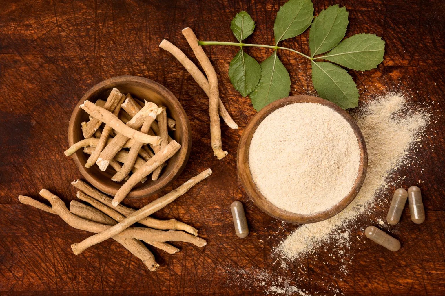 Overhead image of a bowl of ground Ashwaganda powder, and another with root cuttings, and several capsules on a table top