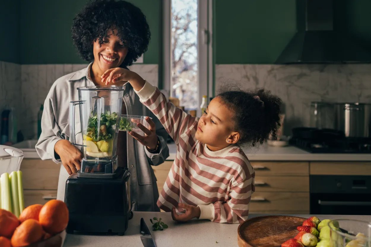 Person and child adding leafy greens and fruit to a blender while preparing a healthy smoothie in a kitchen.