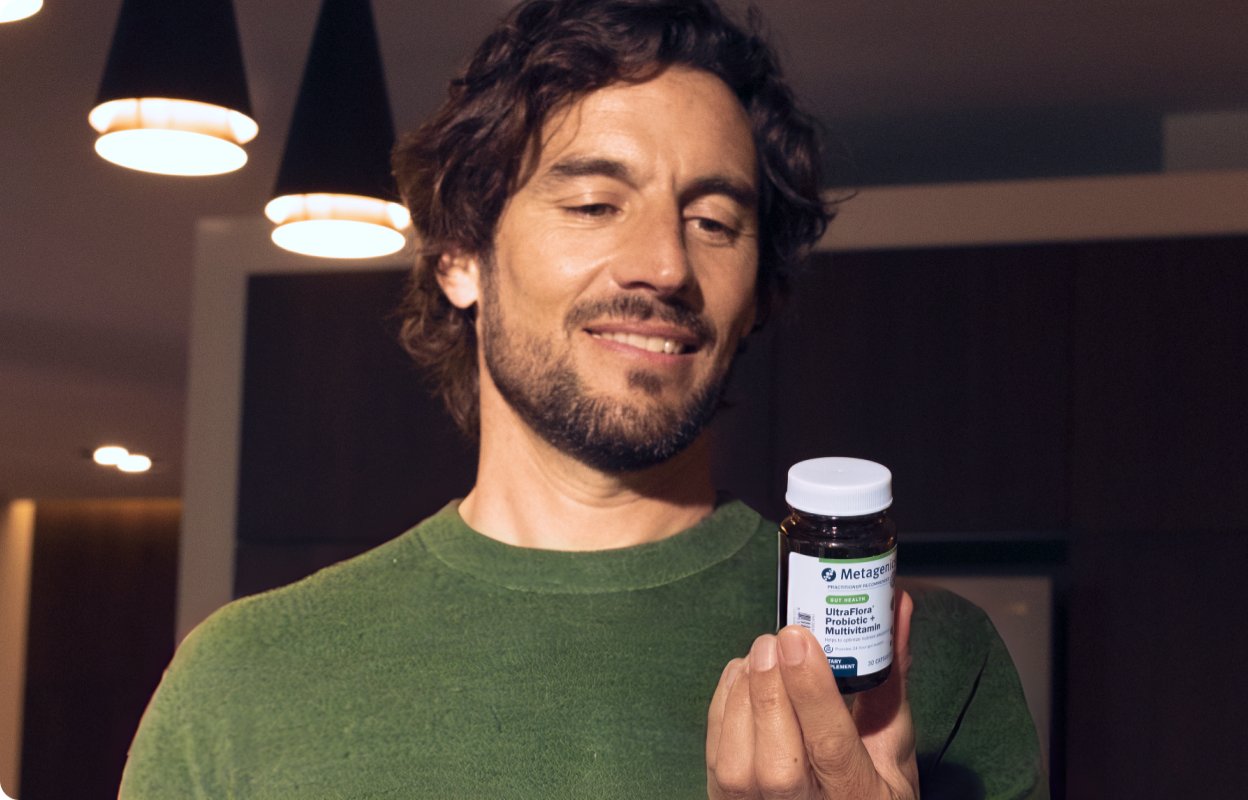 Smiling man with a beard holding a Metagenics UltraFlora Probiotic + Multivitamin bottle in a warmly lit modern room, reading the label.