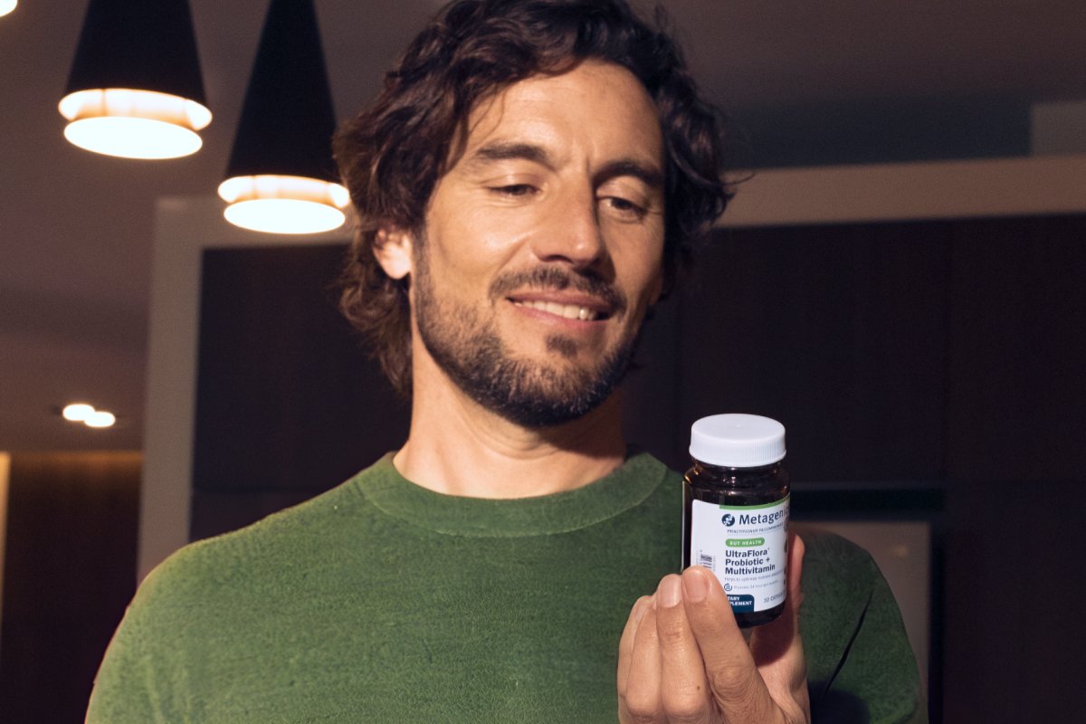 Smiling man with a beard holding a Metagenics UltraFlora Probiotic + Multivitamin bottle in a warmly lit modern room, reading the label.