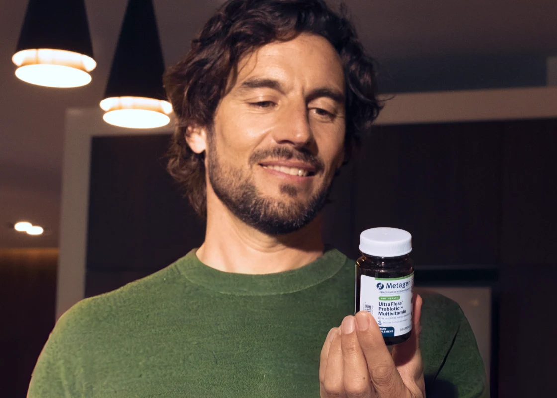 Smiling man with a beard holding a Metagenics UltraFlora Probiotic + Multivitamin bottle in a warmly lit modern room, reading the label.