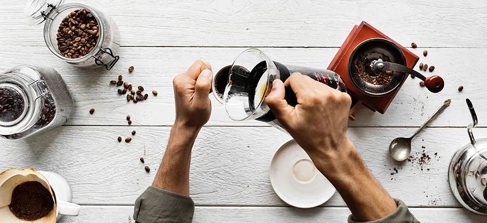 Overhead photograph of a man's arms pouring a cup of drip coffee over a white tabletop with cannisters of coffee beans, filters, and a carafe pot.