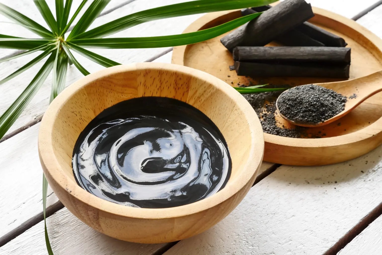 A wooden bowl with a dark liquid adjacent a spoon with crushed charcoal and pieces of charcoal