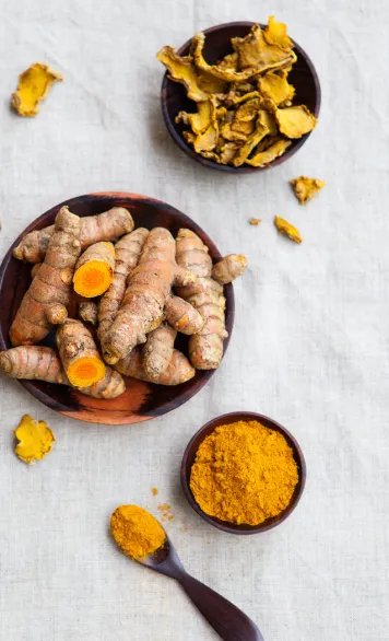 Fresh turmeric roots, dried turmeric pieces, and ground curcumin powder arranged in small bowls with a spoon on a light surface.