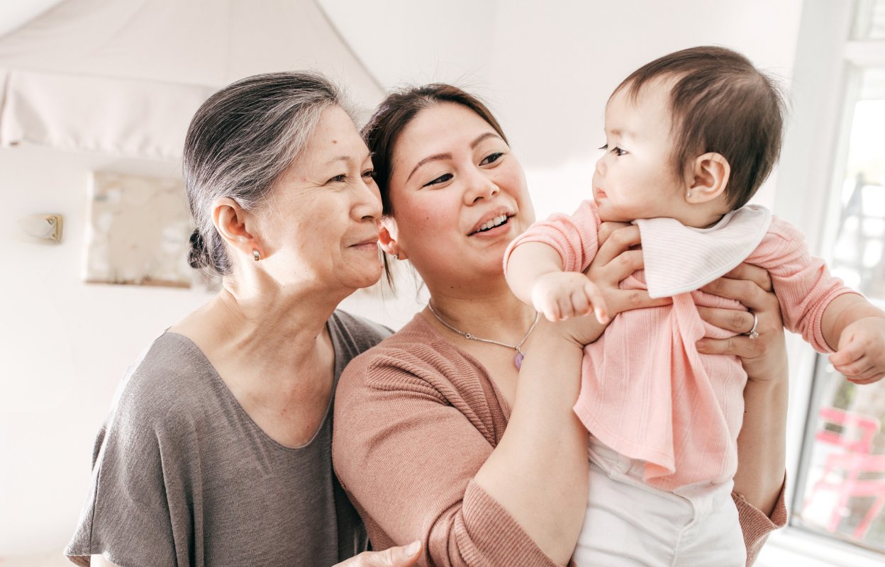 Three generations together in a baby's nursery, a mother holds her infant daughter while grandmother looks on her granddaughter with a smile.