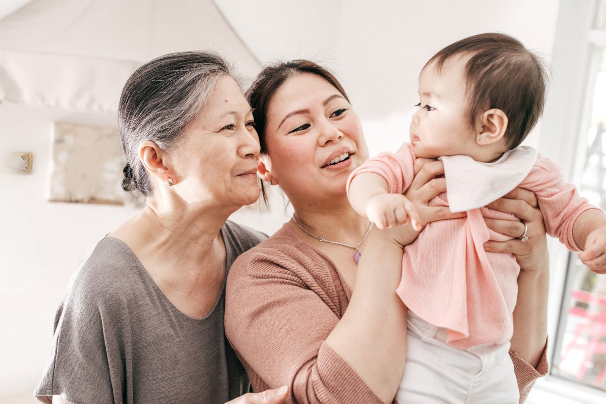 Three generations together in a baby's nursery, a mother holds her infant daughter while grandmother looks on her granddaughter with a smile.