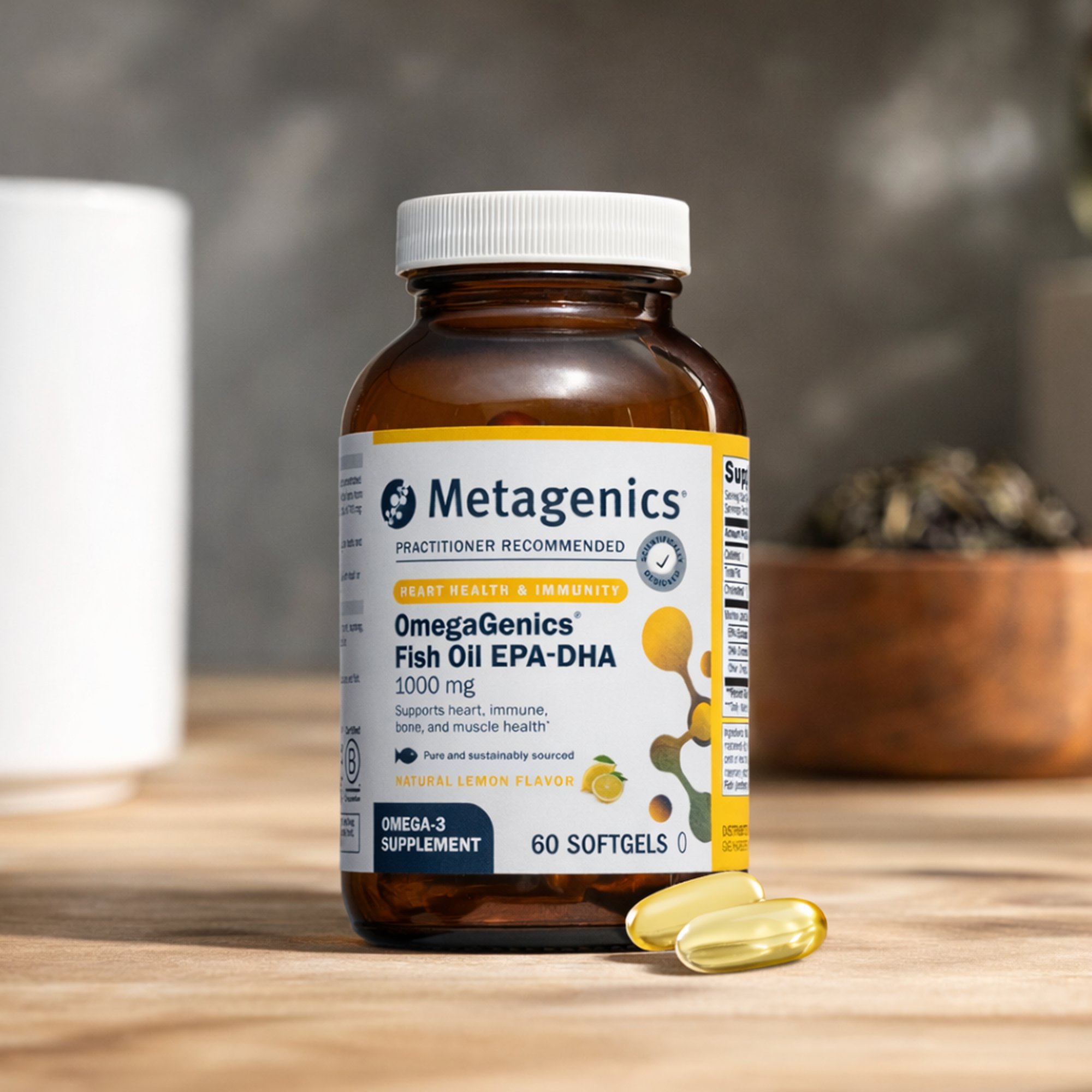 Close-up of a hand holding a bottle of Metagenics OmegaGenics® EPA-DHA fish oil softgels in a bright kitchen setting, with a glass of water nearby. The label highlights omega-3 support for heart, brain, and overall wellness.