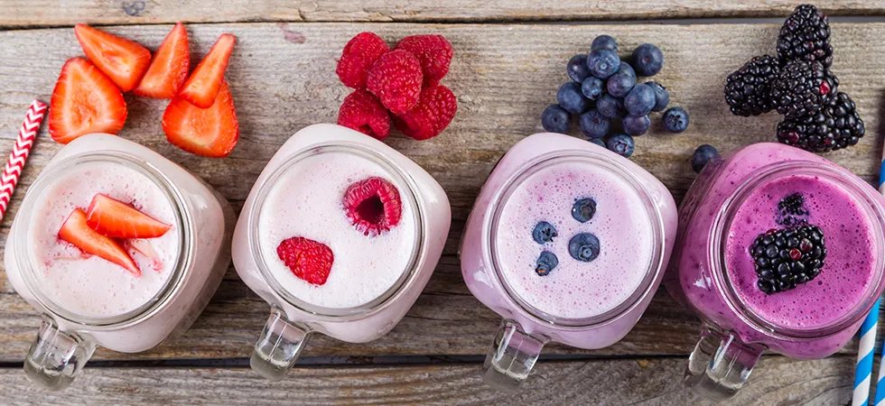 A row of glass mugs with fruit smoothies in each and fresh strawberries, raspberries, blueberries and blackberries next to each one.
