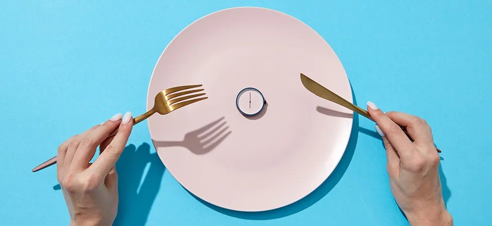 A pink plate with a clock face centered on it, and a woman's hands holding a knife and fork on a blue table top