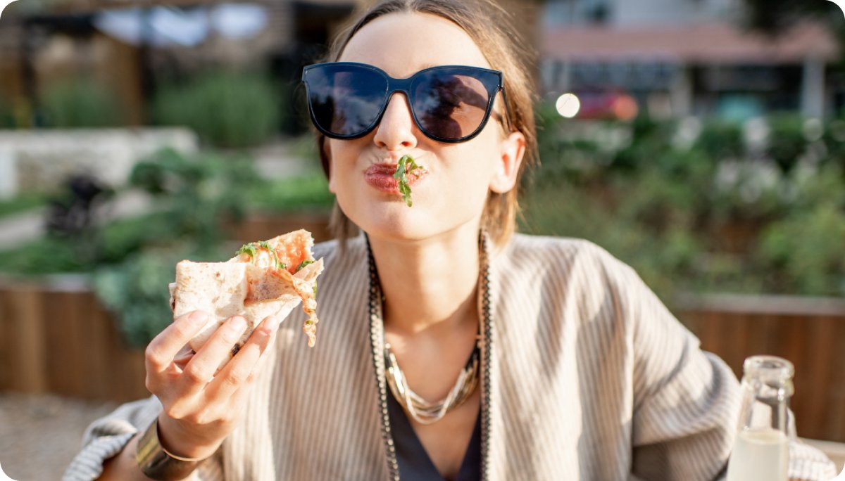  A woman wearing sunglasses eats a slice of pizza outdoors, playfully enjoying food, illustrating the connection between digestion, mood, and the gut-brain axis.