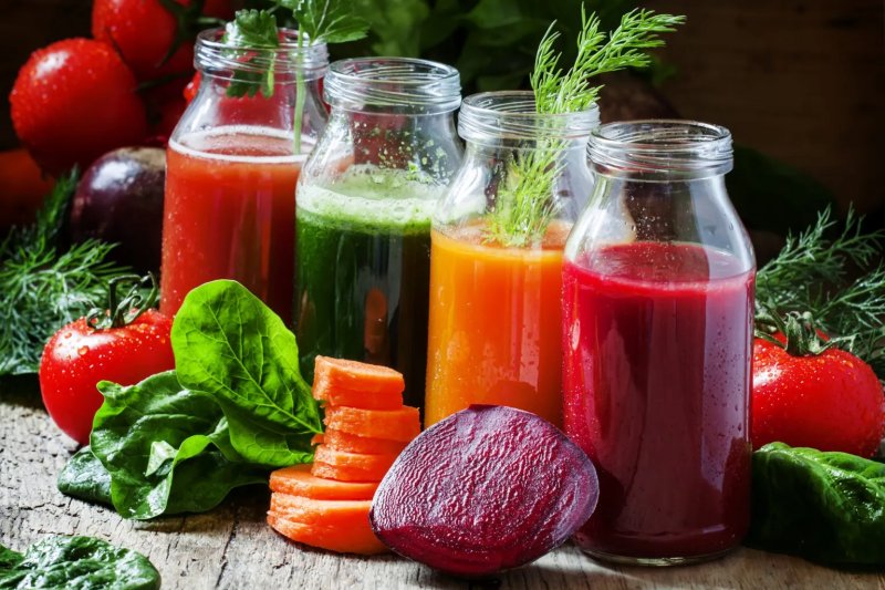 Four bottles containing brightly colored detox juices, and fresh beetroot, carrots, spinach and tomatoes on a table.