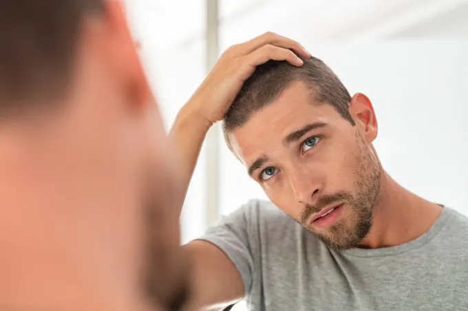Man examining his hair and scalp in a bathroom mirror with his hand resting on top of his head.