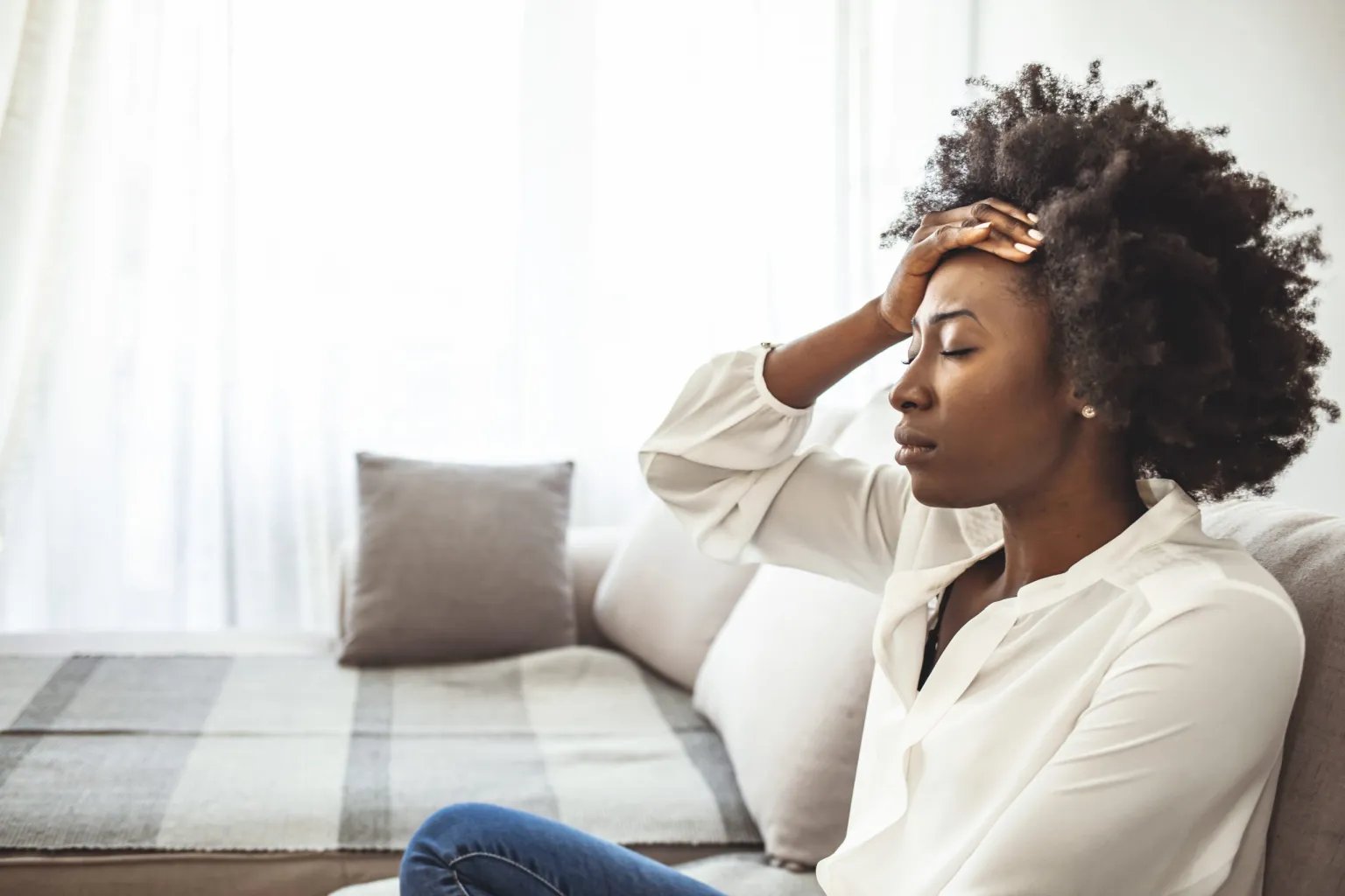 An African American woman sitting on a couch with her hand on her forehead, looking stressed