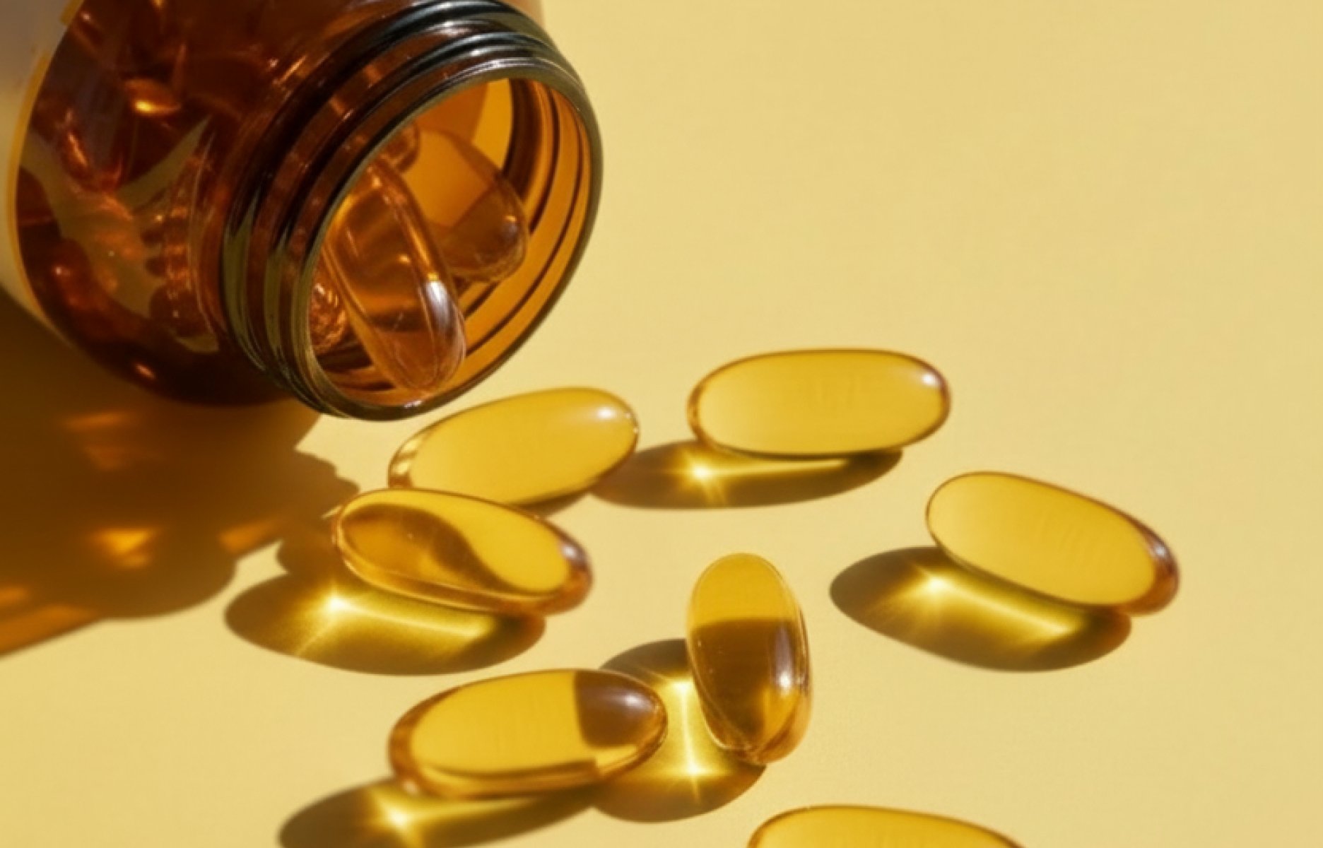 Golden omega-3 fish oil softgel supplements from Metagenics spilling from an amber bottle onto a warm yellow surface.