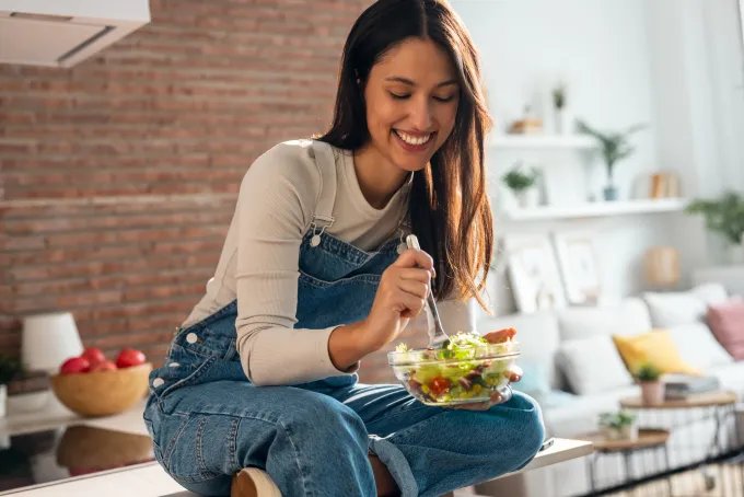 Woman sitting on a kitchen counter and eating a salad from a glass bowl.
