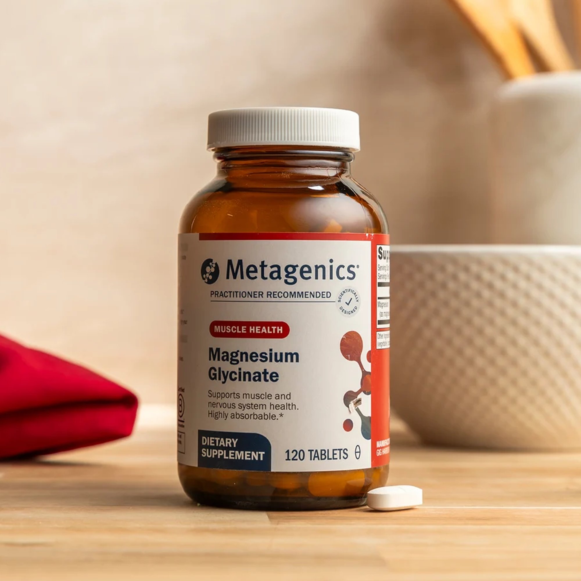 Product image of a Metagenics Magnesium Glycinate dietary supplement bottle placed on a tabletop, with two white tablets in front of the bottle and a ceramic cup and folded red cloth in the background.