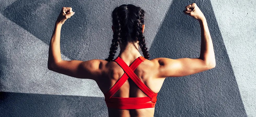 Woman flexing back muscles against a grey wall wearing red sports top